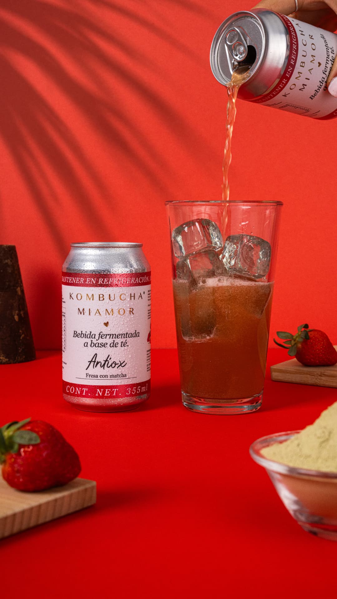 Lata de kombucha Antiox de fresa con matcha servida en vaso con hielo sobre fondo rojo