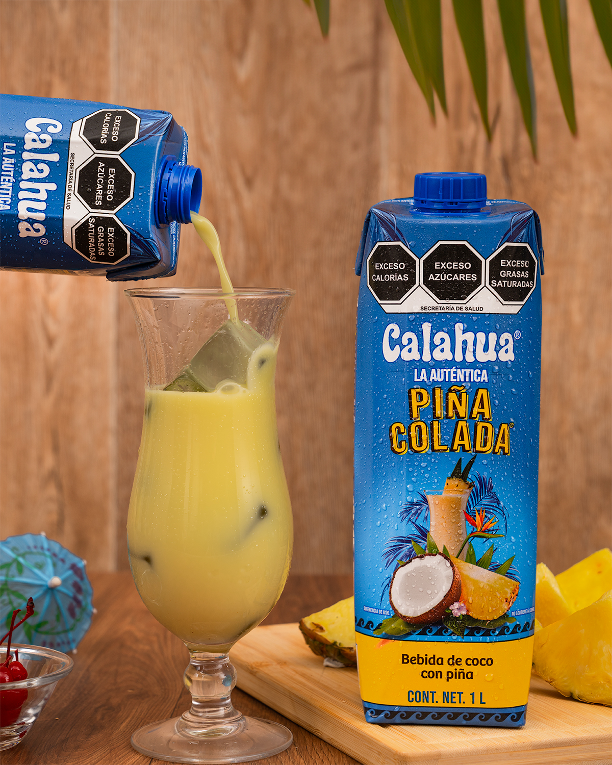 Bebida de piña colada Calahua en envase de cartón sirviéndose en copa con hielo, piña y cerezas alrededor