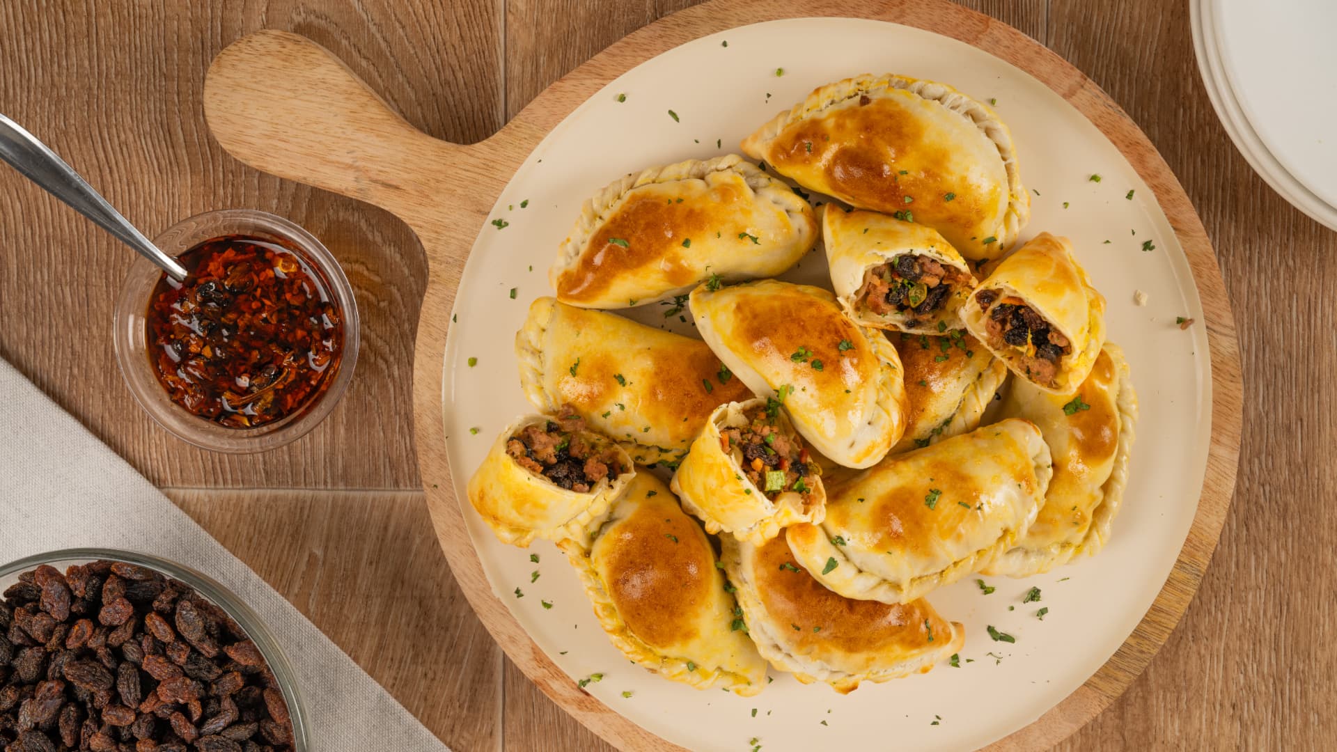 Empanadas de carne con pasitas y hierbas picadas en un plato crema sobre pala de madera, con salsa de chile y un tazón de pasitas, vista cenital