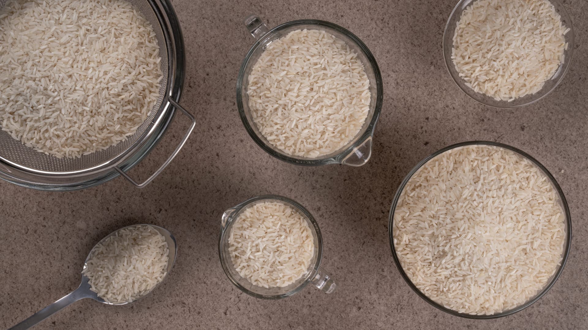 Arroz blanco en colador, tazas medidoras, cuchara y tazones de vidrio, vista cenital
